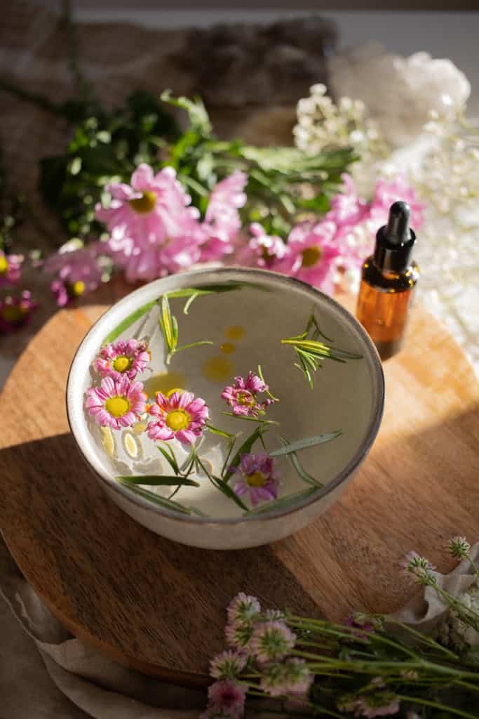 A bowl of water with floating flowers and essential oil bottle on a wooden board in sunlight.