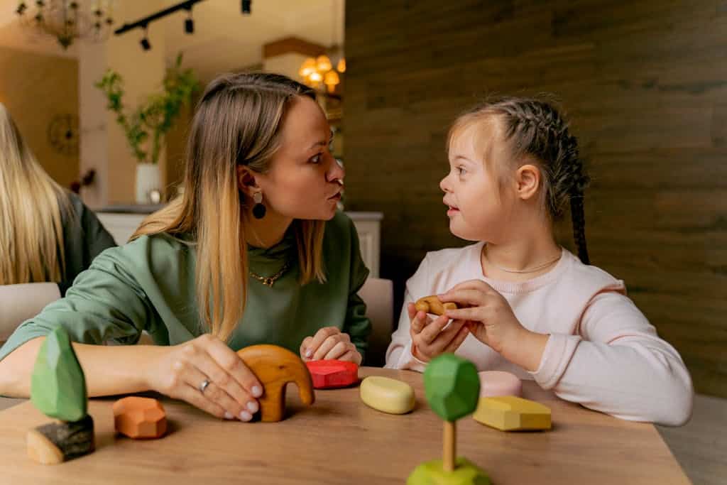 An adult woman and a child with Down syndrome engaging in playful interaction with wooden toys indoors.