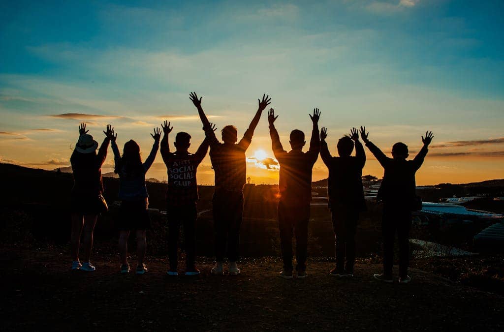 A diverse group of friends raises their arms in celebration against a vibrant sunset backdrop.