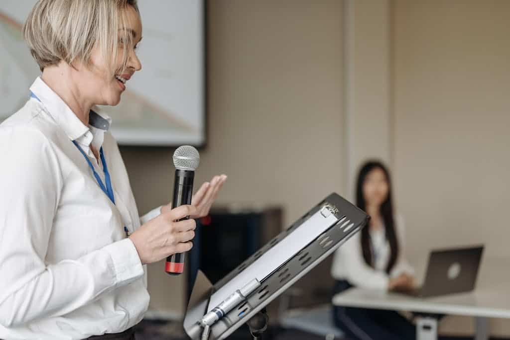 A woman presenting with a microphone at a professional seminar with an audience member in view.