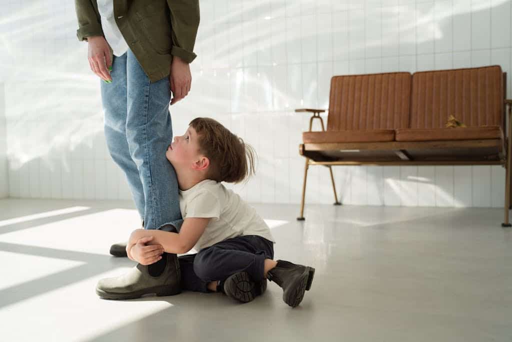 A young boy clings to a parent's leg indoors, expressing emotion in natural light.